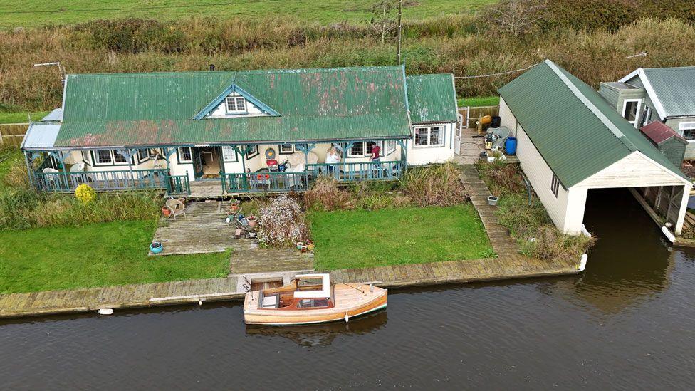 An aerial shot of Towerview. It has a green roof with one triangular dormer window in its middle and cream-coloured walls and is fronted by a veranda. In front of it is a lawn running down to a Broads waterway, where a motorboat is moored. To its right is its green-roofed boat house