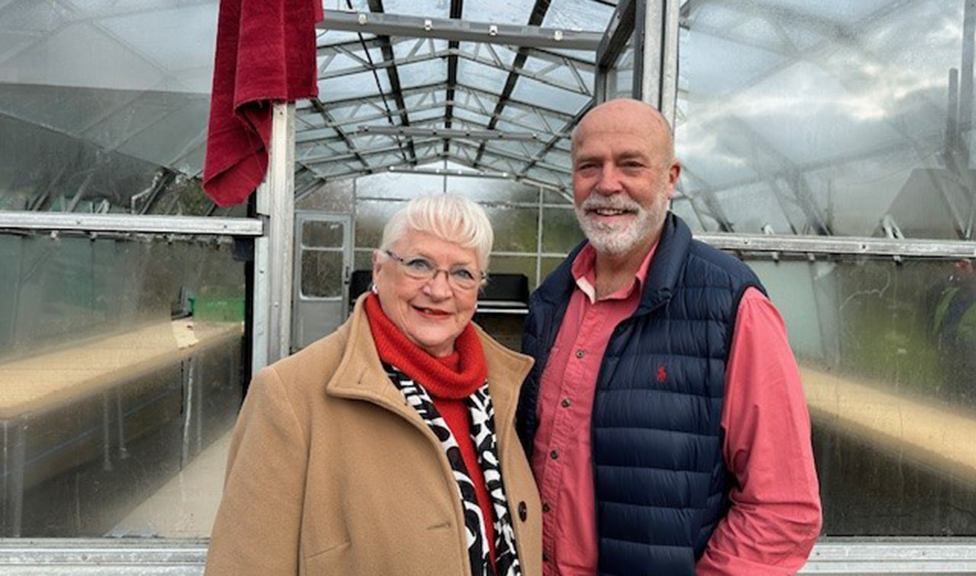 A woman with white hair and glasses in a brown coat and red scarf beside a man with a white beard, red shirt and blue gilet stood in front of a greenhouse