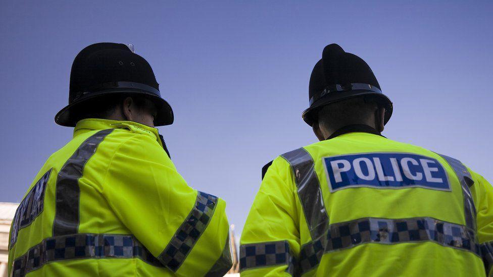 Two police officers in yellow high viz jackets and tall black police helmets stand looking away from the camera on a clear sunny day. 