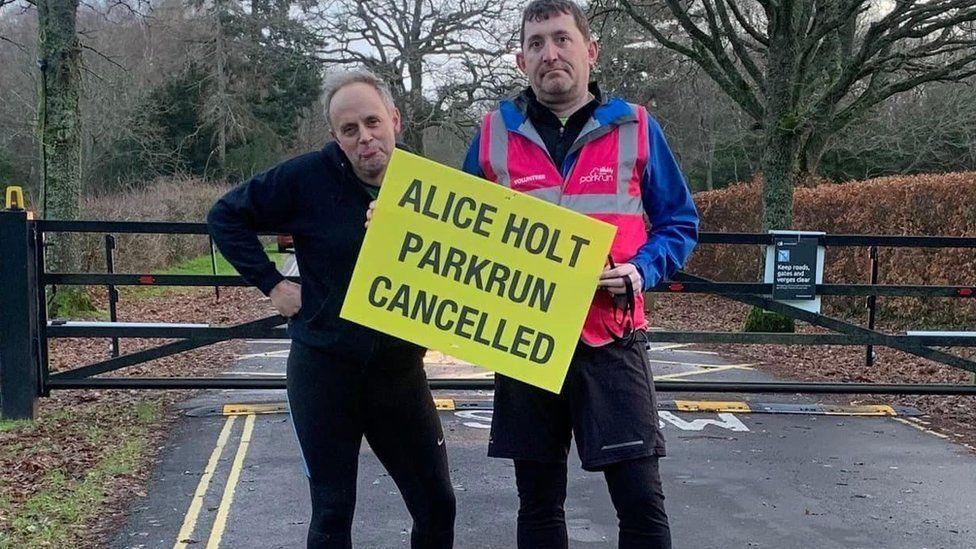 Two men in running clothes standing in front of a metal gate holding a yellow sign with black writing.