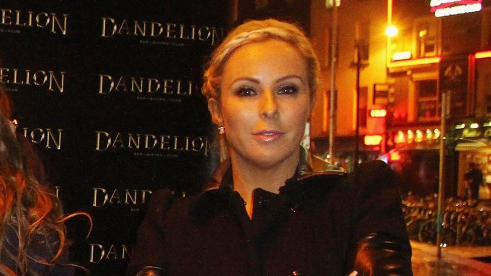 A blonde haired woman with her hair tied back, pictured from the chest up, she is is wearing a black wool and leather jacket and is stood outside at night in Dublin. There is a backdrop advertising the nightclub Dandelion.
