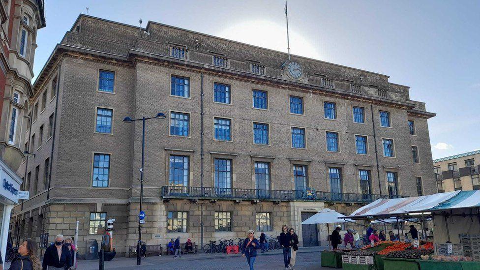 The photo is of the Guildhall but in the bottom right of the photo some stall from Cambridge market can also be seen. The sky is blue and the sun looks to be shining just above the Guildhall building and behind the flag being flow atop it. Just below the flag, on the front of the light bricked building is a clock. There looks to be five-stories to the venue and on the ground floor outside the Guildhall, lots of bikes can be seen parked near to the entrance. 