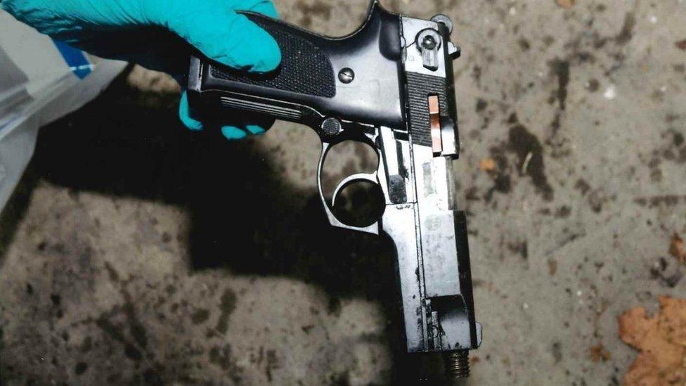 Close up of a forensic officer, wearing a white hazmat suit and blue latex gloves, holding a black pistol by the handle with the barrel of the weapon pointing at the ground. 