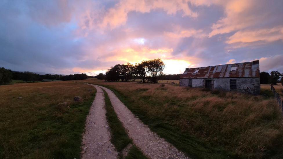 An old farm building next to a track at sunset