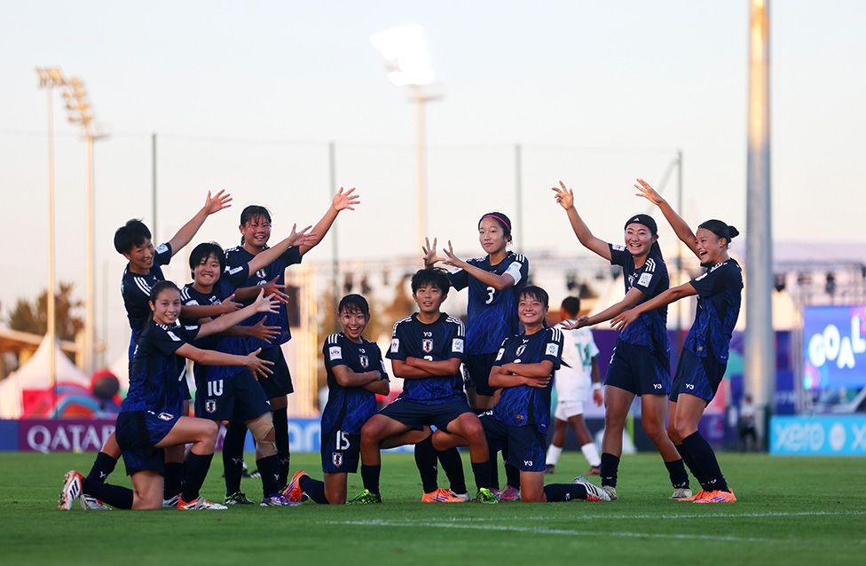 A women's football team in dark blue uniforms kneeling and standing on a field, posing with arms raised in various directions during daylight, with stadium lights and banners visible in the background.