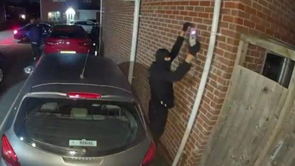 A CCTV image showing a person dressed in black clothing and wearing a balaclava holding a device against a brick wall. The individual is standing in a residential driveway next to a silver car parked close to the wall, with other vehicles visible in the background under street lighting. A wooden fence is partially visible on the right side.