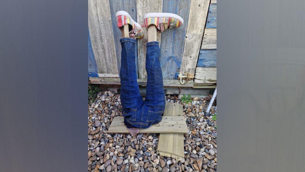 A pair of fake legs are placed against a garden shed. They are made from jeans and wooden poles and there are a pair of colourful shoes on the wooden feet.