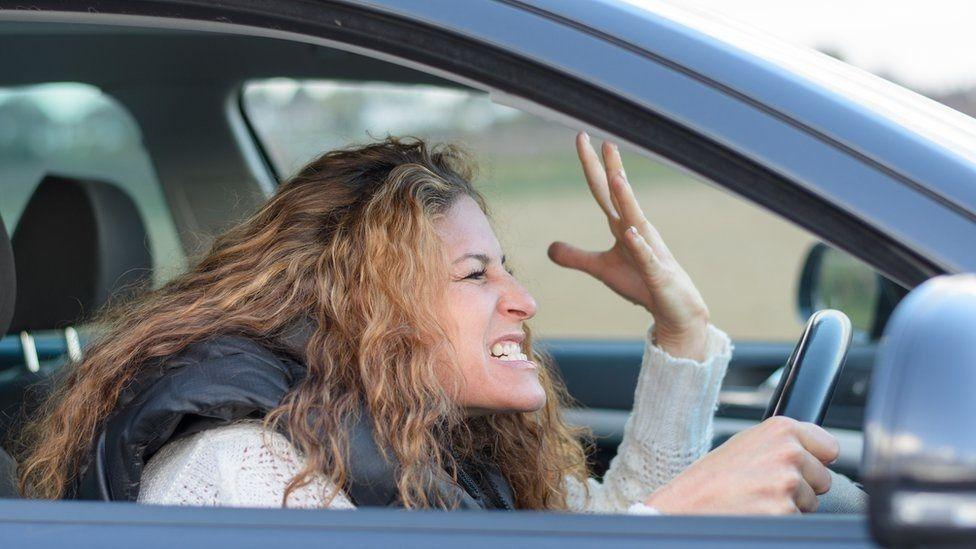 File picture of woman with long brown hair with an angry face, gritted teeth and a left hand outstretched. She is wearing a white top and black gilet and sitting in the driver's seat of a grey car.