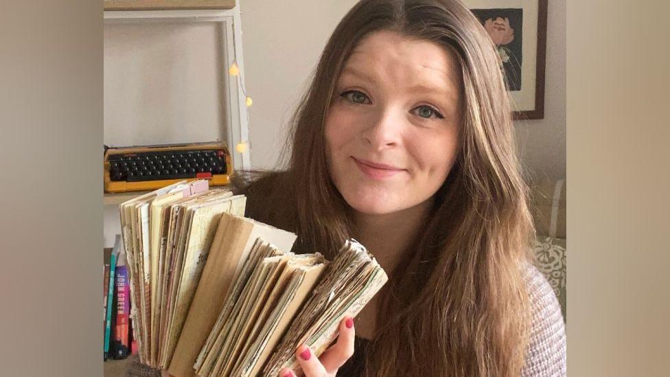 Meg has long brown hair and is holding a stack of brown paper journals. She is sitting in a room with cream coloured walls and fairy lights