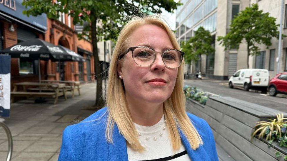 Nicola Armstrong, with blonde hair and glasses, wearing blue jacket, white and blue striped jumper and beaded necklace.