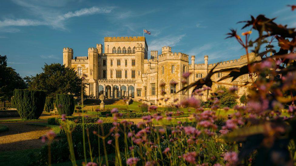 A castellated stone manor house against a blue sky with yew trees, box hedging and pink flowers in front, Ashridge House 