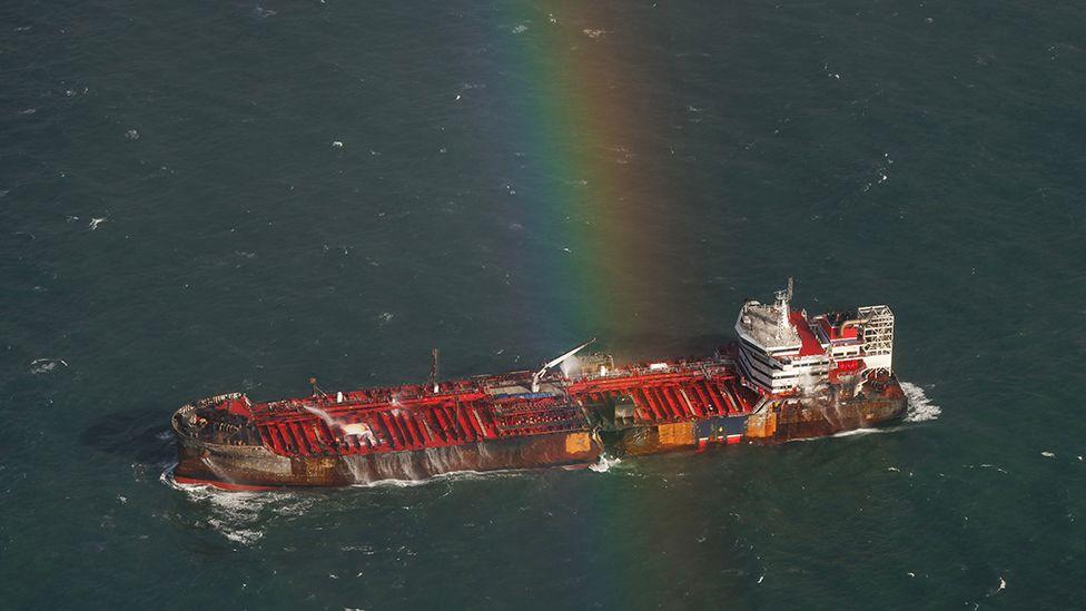 Aerial view of the Stena Immaculate showing a hole in the hull of its left side signs of fire damage to the hull and a rainbow above the ship. 