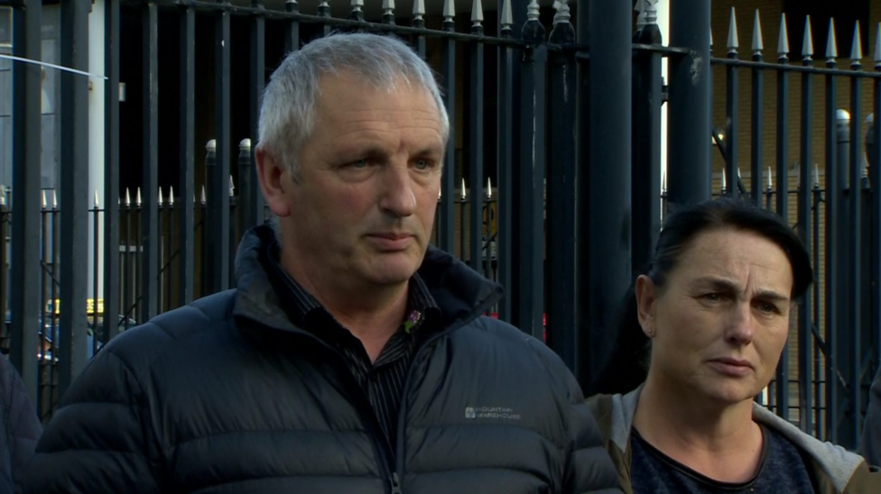 Michael McConville is standing looking slightly off camera with his lips pursed. He is wearing a black coat and black shirt. To his left a woman stands looking on. Behind them are black railings.
