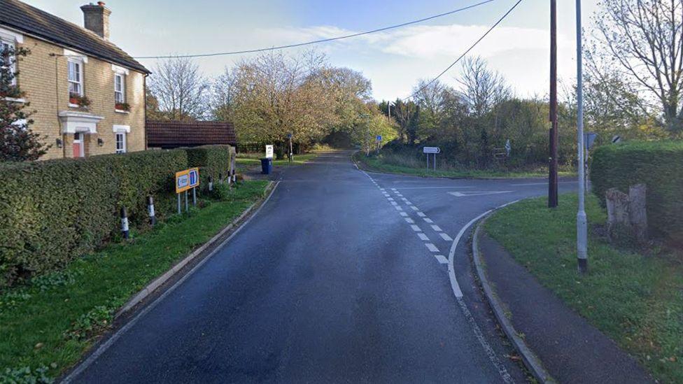 A view of the junction from Hilton Road into Connington. There is a house on the left side, behind a hedge, and a road sign to Connington and Fenstanton next to the junction. There are telegraph wires overhead and two posts on the right. Trees and fields are in the distance.