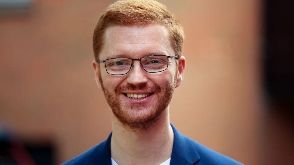 Ross Greer, a ginger man with glasses, smiles at the camera. He is wearing a blue jacket and white t-shirt.
