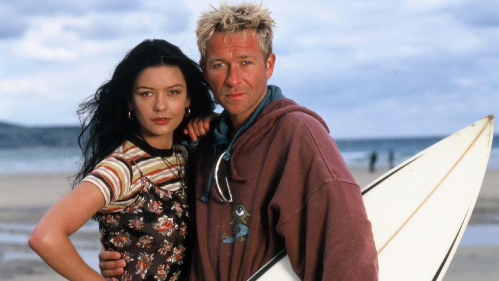 A young Catherine Zeta-Jones, who is wearing a flowery dress over the top of a striped T-Shirt, posing with Sean Pertwee, who has a surfboard under his arm, on a beach.