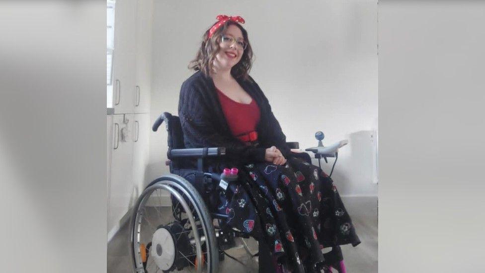 A woman with dark hair with a red ribbon in it, wearing a red top and a black patterned skirt, sitting in a wheelchair and smiling at the camera.