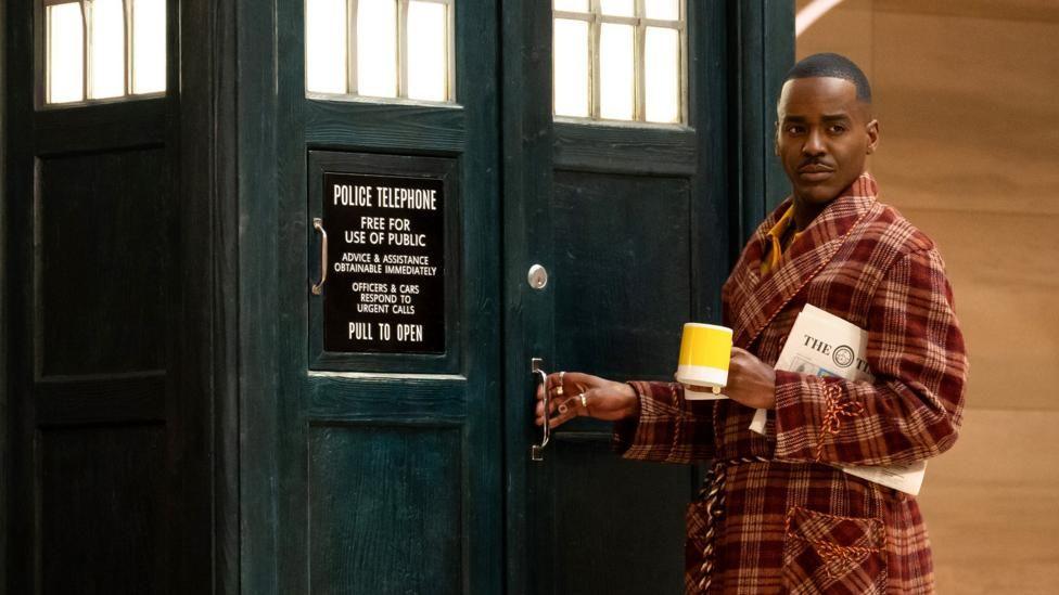 Ncuti Gatwa as the Doctor in a dressing gown holding a mug of tea and a newspaper. 