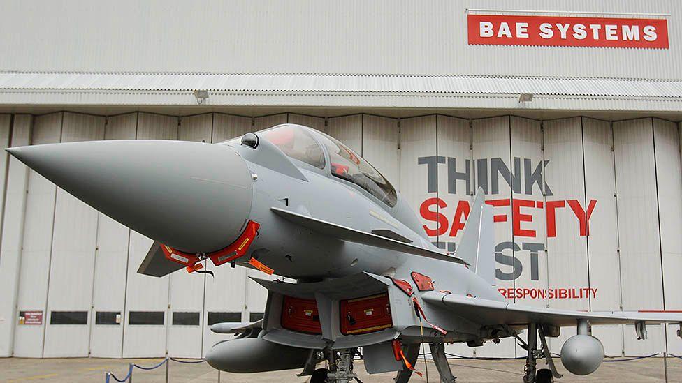 A Eurofighter Typhoon fighter jet at BAE Systems in Warton stands outside a grey warehouse with concertina doors and a red-and-white BAE Systems sign.