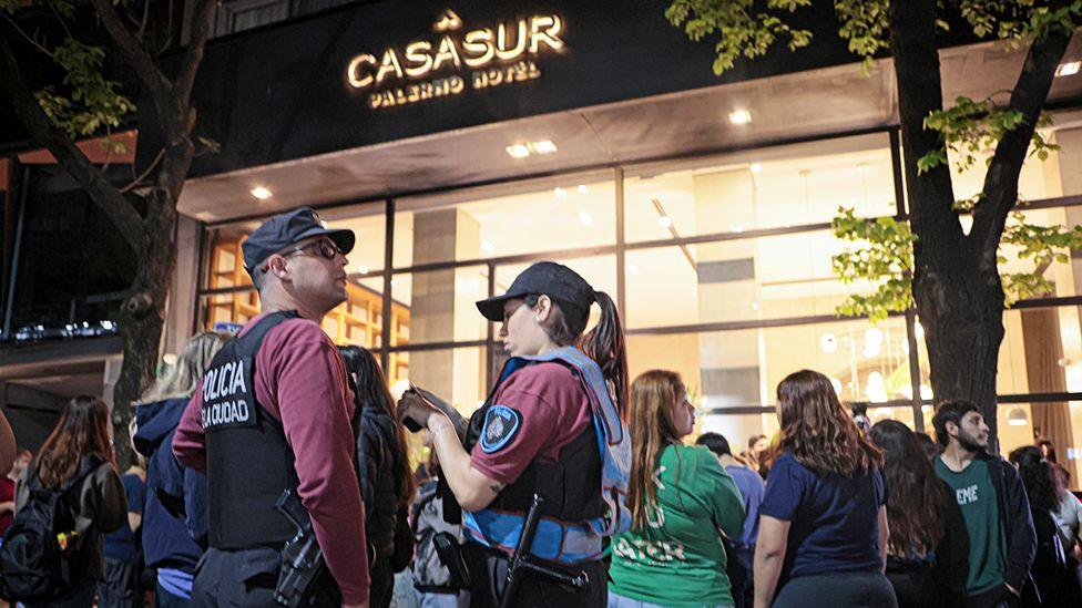 Police officers in tac gear saying 'Policia' on the back and wearing police caps stand in front of a crowd of people outside the CasaSur Palermo hotel in Buenos Aires, with the hotel's sign visible in the background, on 16 October