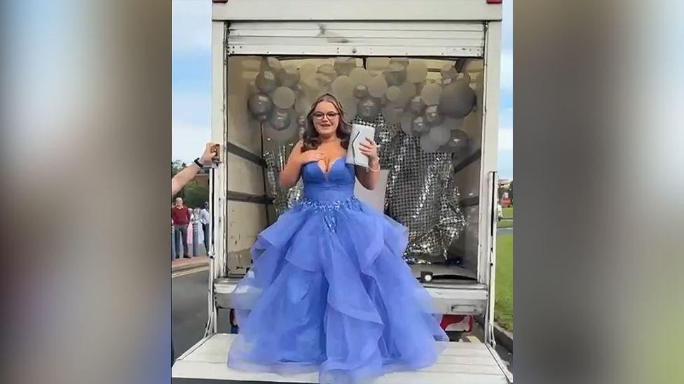 Jessica, with long dark hair and black glasses, wears a purple prom dress as she stands on a tail lift on the back of a removals van. In one hand she is holding a white handbag.