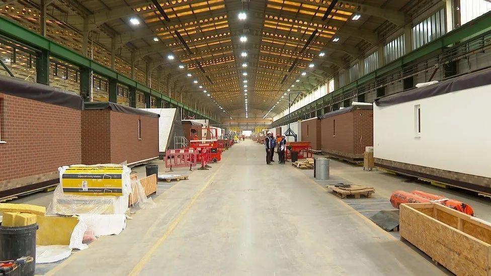 Pre-fabricated home sections on with sides of a corridor in a large warehouse. 