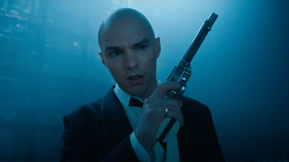 A bald Nicholas Hoult in a black suit holding a gun in a still from the trailer