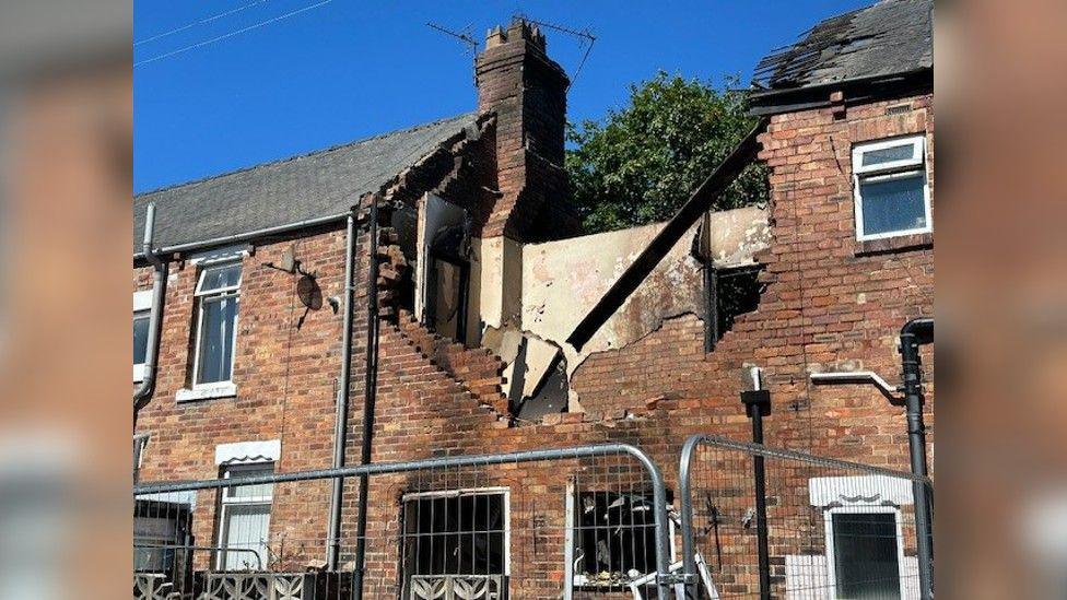 The damaged house, with a fence around it, with its roof destroyed.