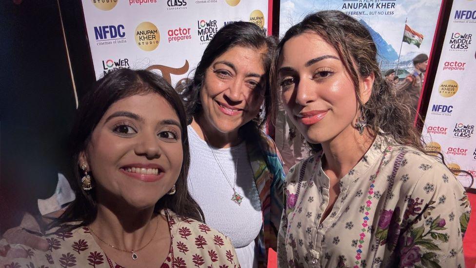 Three woman standing on a red carpet smile in a selfie. In the background is a large white board with company logos on it