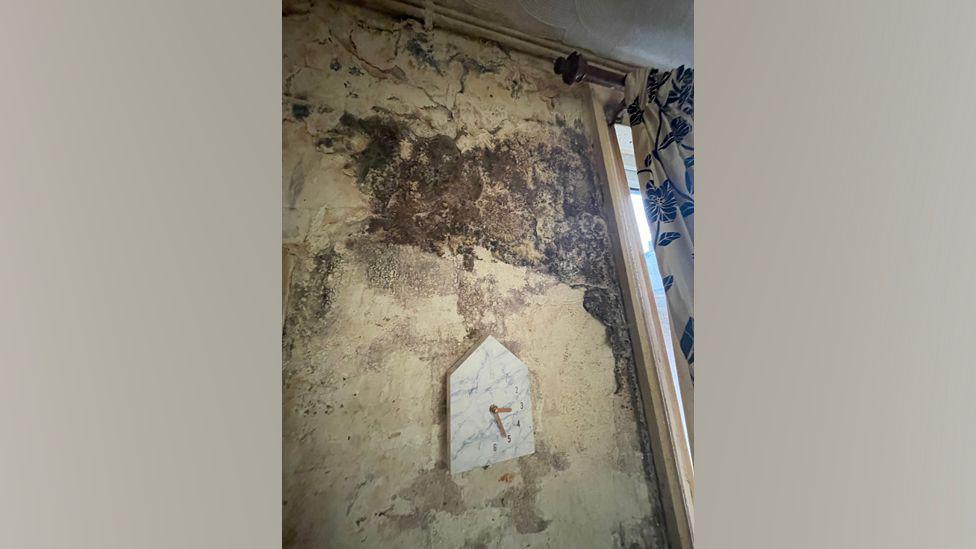 A clock hanging on a dirty wall inside one of the homes. Black mould is growing across the bare wall. A pair of curtains is hanging by a window to the right.