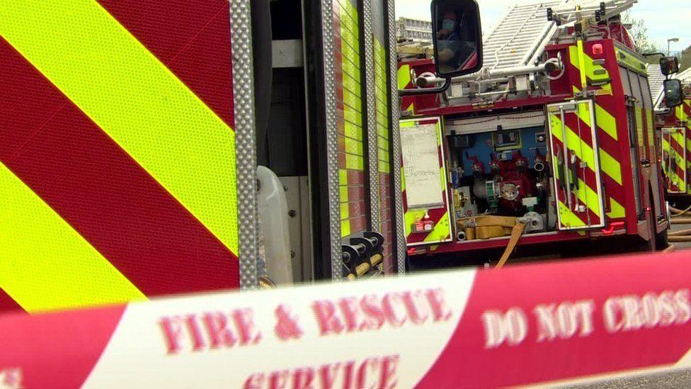Two fire engines are shown close up and red and white tape cordons off a scene.