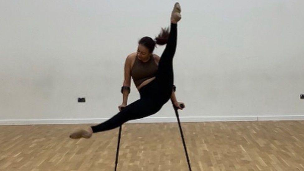 In a studio with a wooden floor Sophia Adzoa-Moore is doing the splits in the air using a pair of crutches.