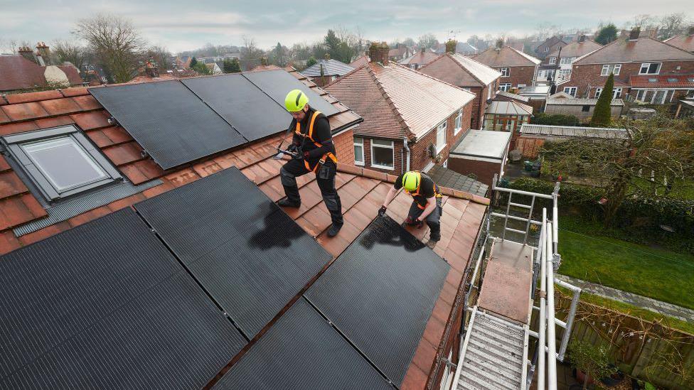 Workmen on the roof of a house installing solar panels while wearing orange harnesses and yellow helmets 