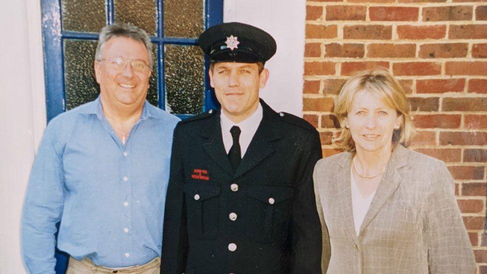 Jonathan Rudd in his navy blue firefighter jacket and hat with his father Nigel to his left in a light blue shirt and his mother to his right in a beige checked suit jacket