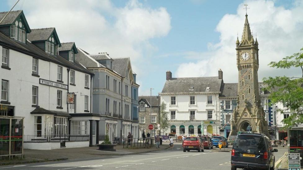 A main street in Machynlleth leading to a tall stone clock tower in a square and townhouses around the edges. There are cars on the road. It is a blue sky with some clouds.
