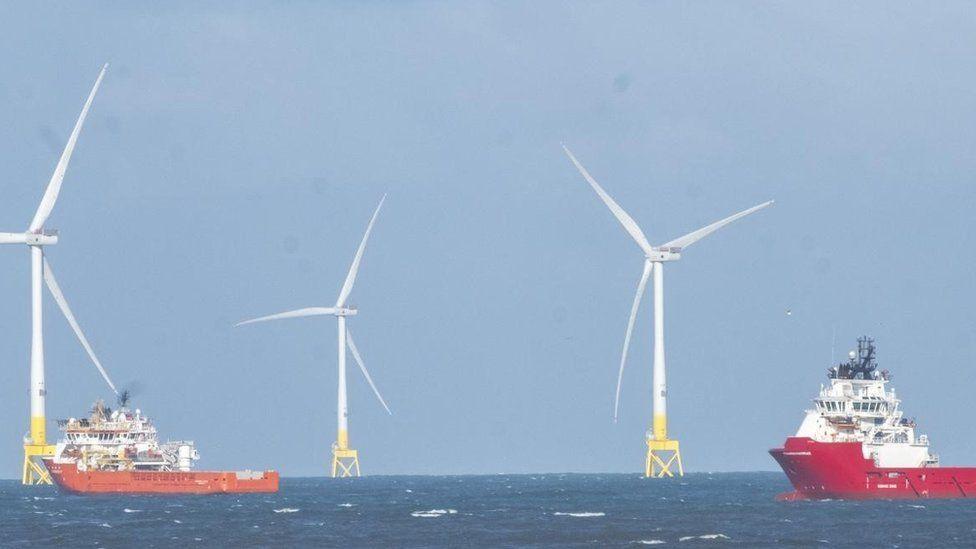 Sea with two red and white boats and large white wind turbines on yellow bases towering above.