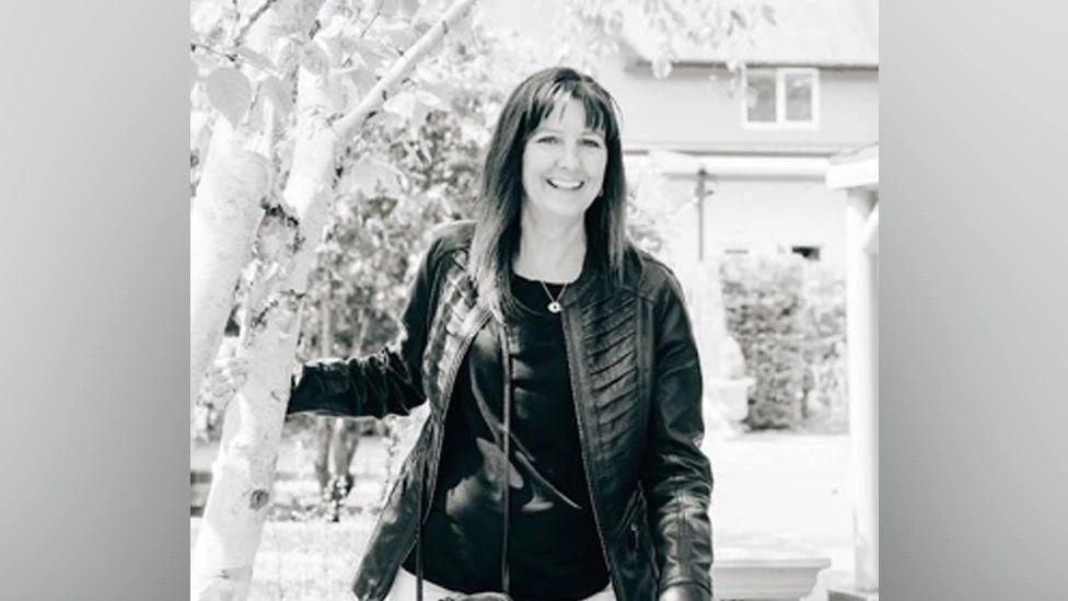 A black and white image of a smiling woman with shoulder-length hair wearing a leather jacket and necklace