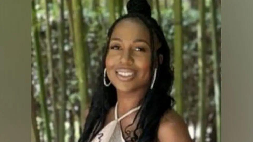 Michelle Sadio smiles at camera wearing large white hooped earrings and a pale coloured dress, in front of green bamboo plants