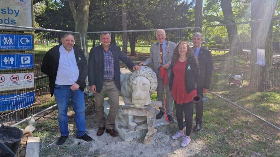 A group of people looking at the camera while stood around the restored lion the area is fenced off and trees and grassland can be seen in the background