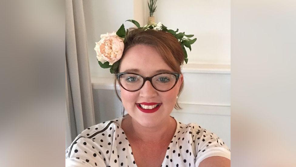A photo of a smiling woman who is wearing a white t-shirt with black polka dots, black framed glasses, bright red lipstick and a headband with flowers on. She is photographing herself from the shoulder up.