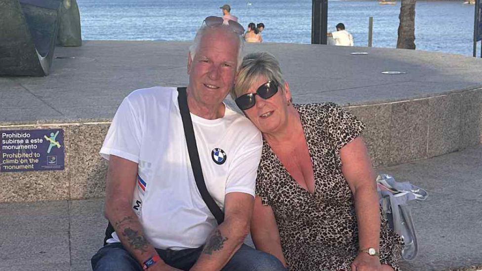 Alan is sat on the left of the image, wearing a white T-shirt and a cross-body bag. His partner is sat on the right, in a leopard print dress, with her head on his shoulder. Behind them is a large body of water.