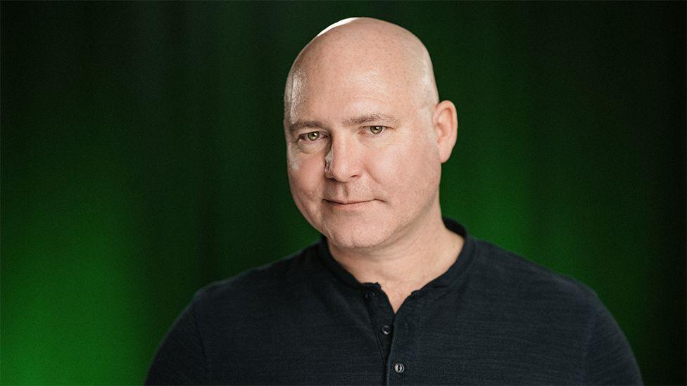 The voice over actor, Doug Cockle, who is a bald man - smirking with a black buttoned top in front of a black and green background. 