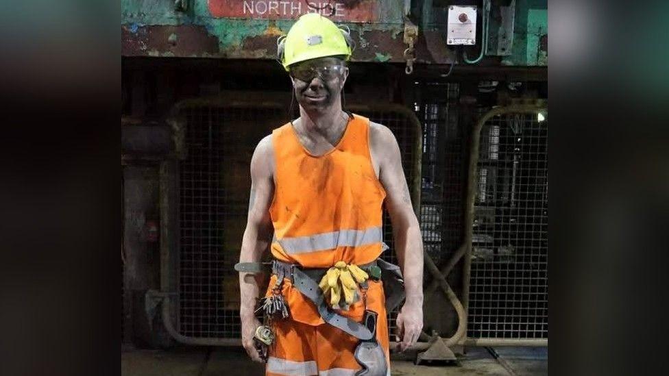 Shane Harrison wears a yellow hard had, clear glasses and orange hi-vis clothing. He also has a utility belt with keys and gloves attached. His face is dirty from being down the mine, and he is stood in front of machinery.