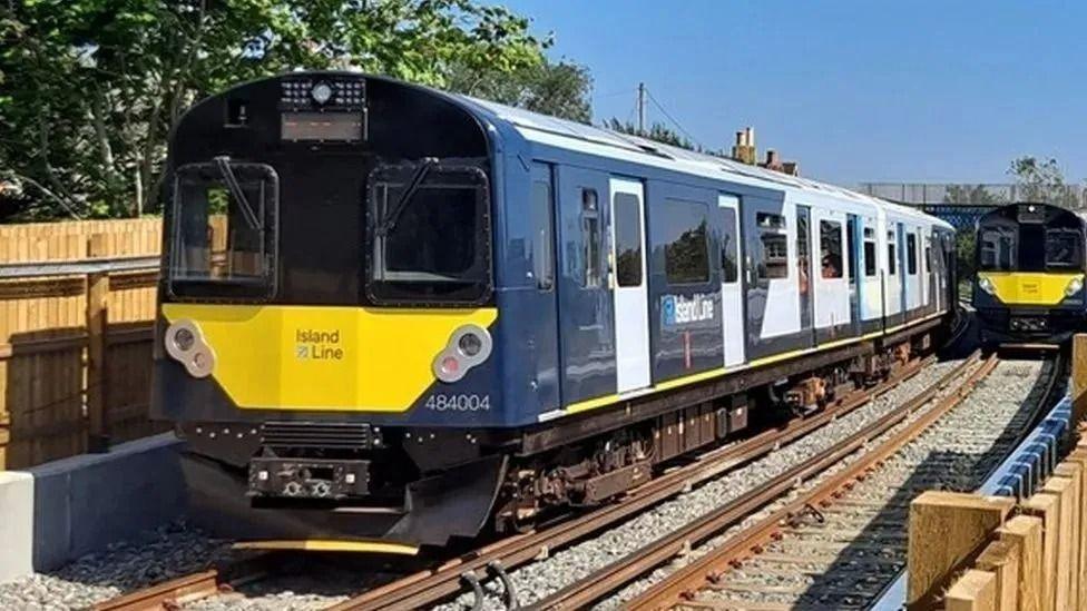 A Island Line train on the track. It is blue, yellow and white. A second identical train can be seen in the background.