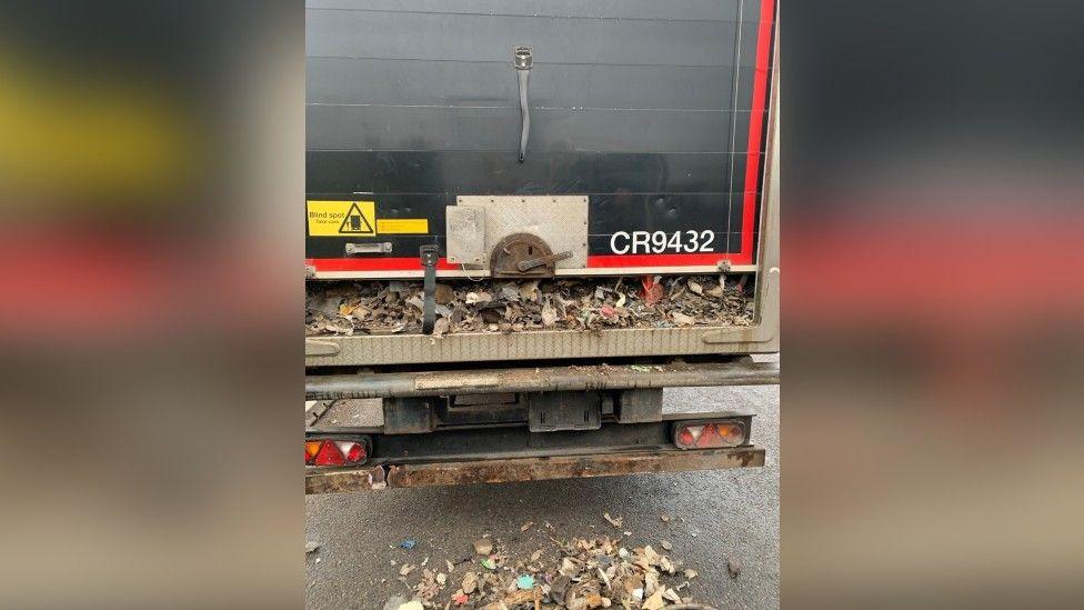 Rear of a trailer with the roller door slightly open, allowing some of the fist-sized pieces of mixed, dry waste to spill out on to the road