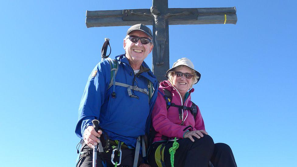 Ray Craven and his wife Janice, wearing climbing gear, smiling, next to a large crucifix statue, clearly high up with a blue sky behind them. 