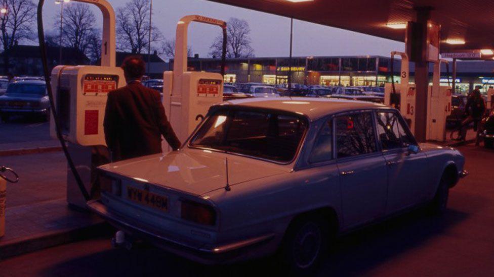 A back view of a man in a jacket standing behind a car and in front of a white petrol pump. He is filling the white car with his right hand. It is dusk and beyond the forecourt can be seen a Sainsbury's store and leafless trees framed against the sky