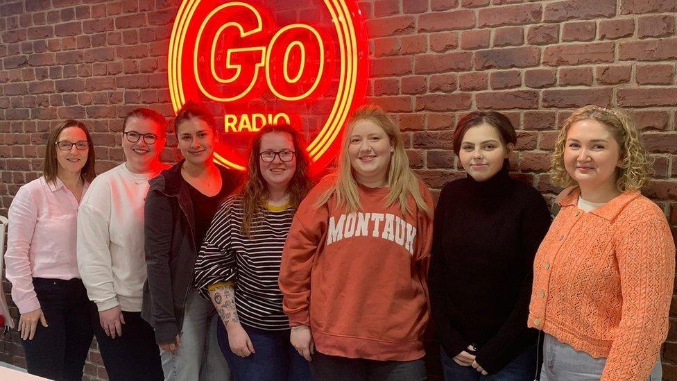 Molly (second right) with six other members of the Go Radio podcast team - they stand in a line in front of a brick wall with a Go Radio neon sign.