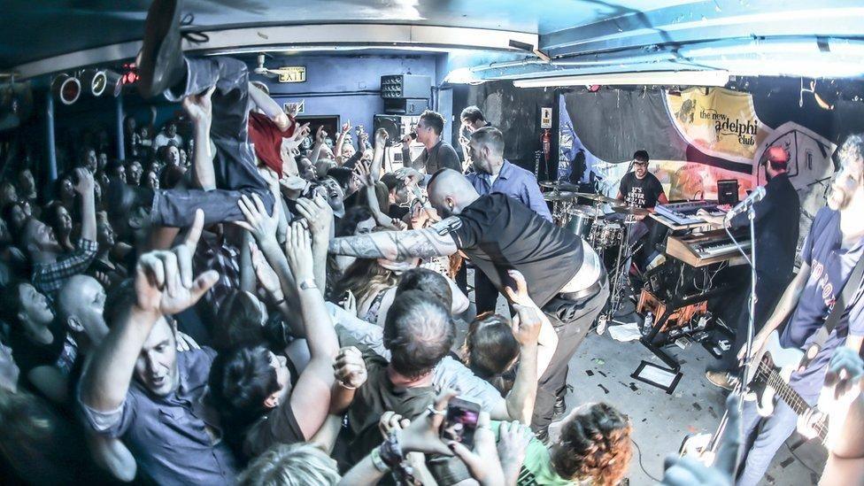 A crowd of revellers - men and women - going wild with one individual crowd-surfing above them towards the front of the stage where six musicians are performing. A security guard is pushing the crowd-surfer back  away from the stage 