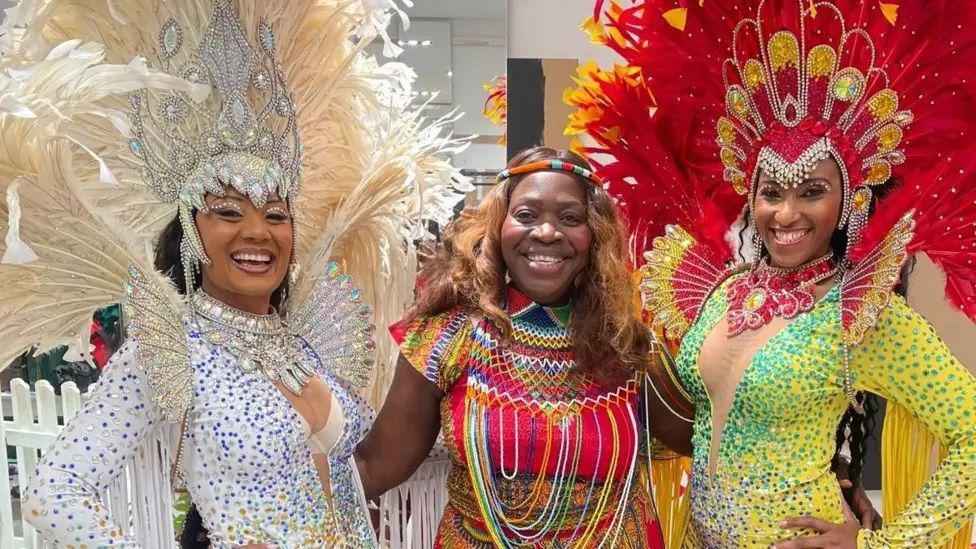 Three women including Bernadetta dressed in colourful Caribbean dance outfits. Bernadetta has her arms around both of them. The two women on either side both have sequin dresses and feathered headgear. The one on the left is in a white and blue outfit. The one on the right is in a yellow and emerald outfit, with a red headdress.